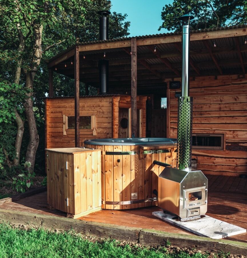 Escape to Woodland Chase, where a rustic wooden cabin awaits you. Nestled in a lush forested area, it features an inviting outdoor hot tub and cozy stove amidst towering trees and vibrant green grass—a perfect retreat into nature's embrace.