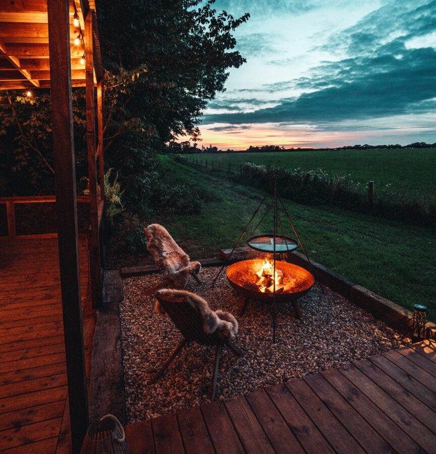 A cozy outdoor seating area with a fire pit and two chairs, nestled among woodland greenery, under a partly cloudy evening sky.