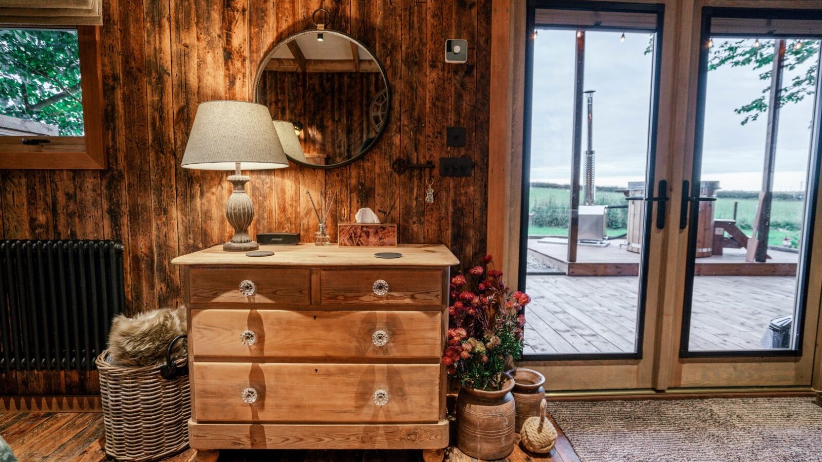 Experience the charm of Woodland Chase Cabins, with its cozy wooden cabin interiors. Featuring a dresser adorned with a lamp, round mirror, and plants, this serene space opens to a rustic outdoor view that invites you into the tranquil embrace of the woodland.