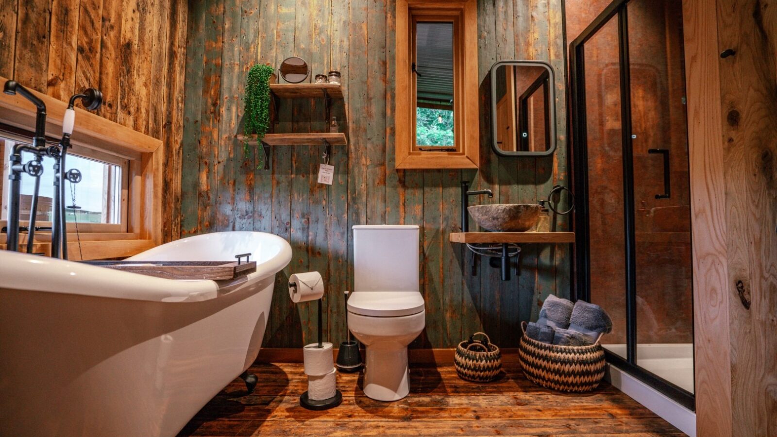 Rustic bathroom reminiscent of woodland cabins, featuring a freestanding bathtub, wooden walls and floor, toilet, shower with glass door, and woven baskets.