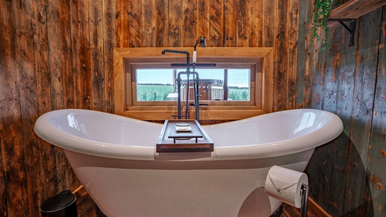 Freestanding bathtub with a tray, nestled in a rustic bathroom with wooden walls and a window view of the lush Woodland Chase.