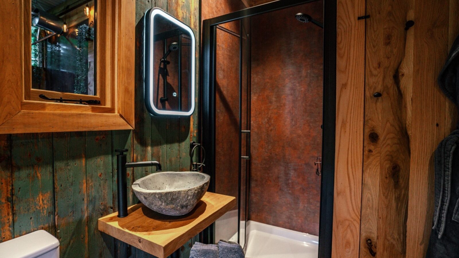 A rustic woodland bathroom features a stone sink on a wooden counter, lit mirror, and a glass shower with a dark metal frame surrounded by wood-paneled walls. Perfect for those who chase the tranquility of nature.