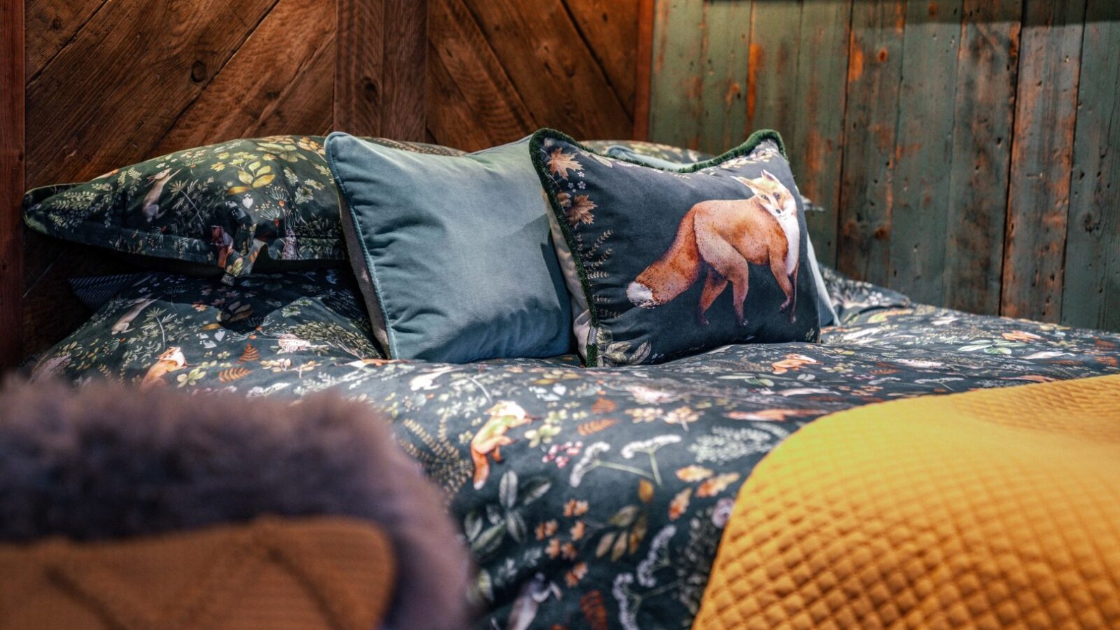 A rustic bed with fox-themed pillows and a forest-patterned duvet evokes the essence of Woodland Chase in a cozy wooden room, complemented by a yellow blanket at the foot—ideal for cabin lovers.