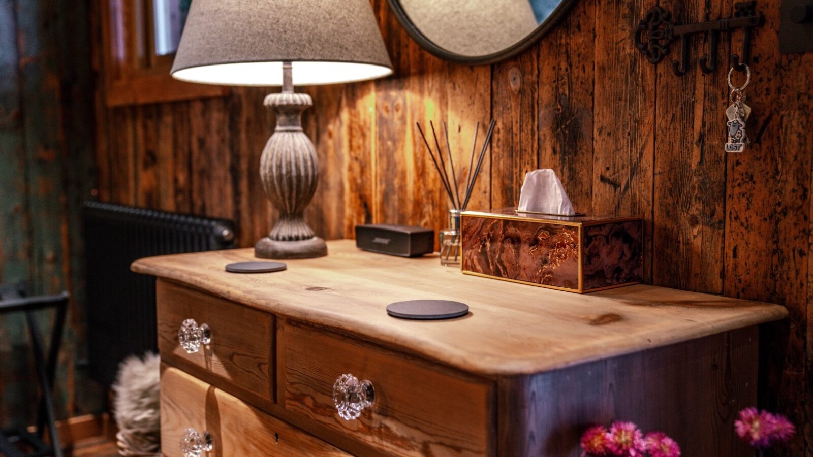 A wooden dresser adorned with a lamp, round mirror, tissue box, and keys sits against a rustic wall reminiscent of the Woodland Chase Cabins. Surrounded by flowers, this charming scene invites a cozy escape.