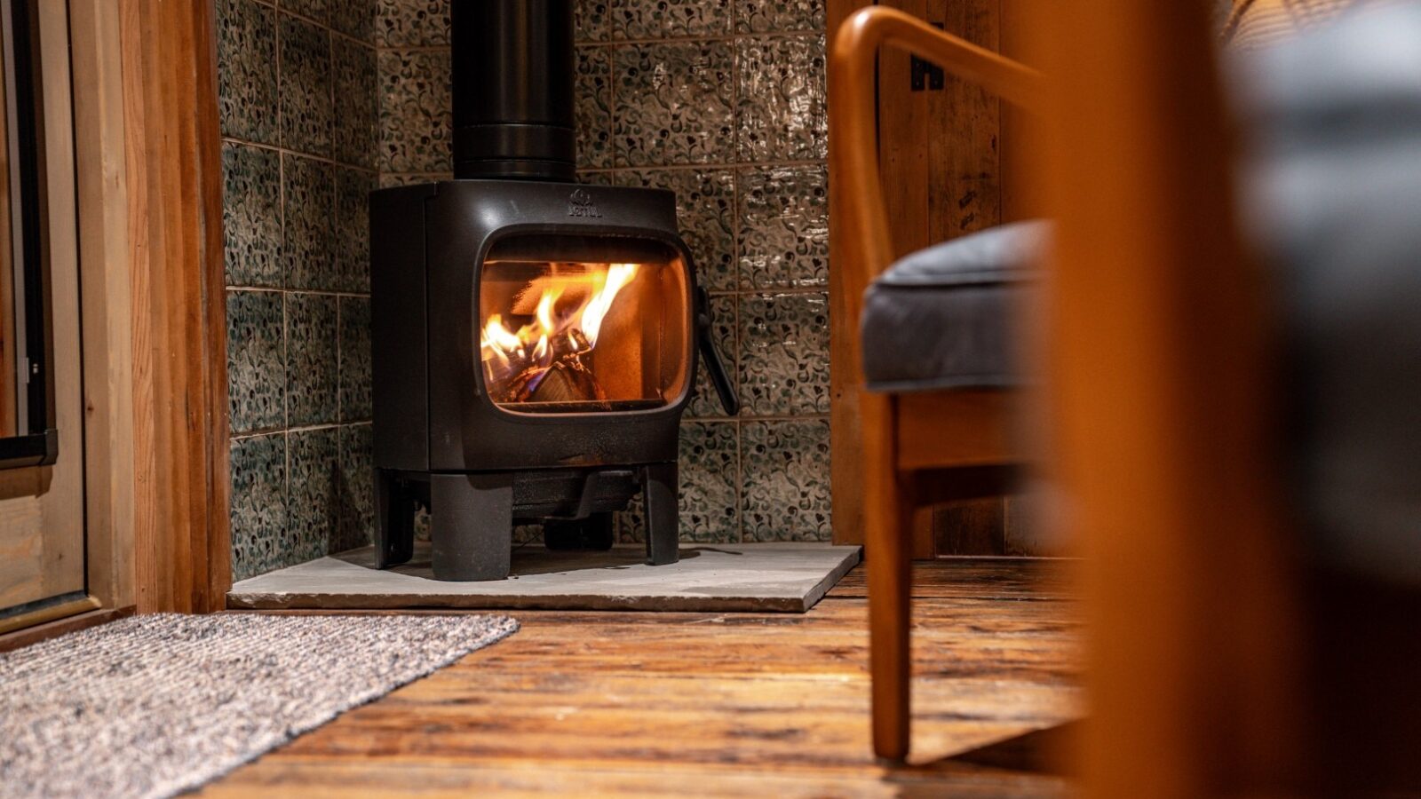 A cozy room reminiscent of woodland cabins, with a lit wood-burning stove, wooden floors, and a patterned tile wall. A partially visible chair sits in the foreground, inviting you to chase away the chill.