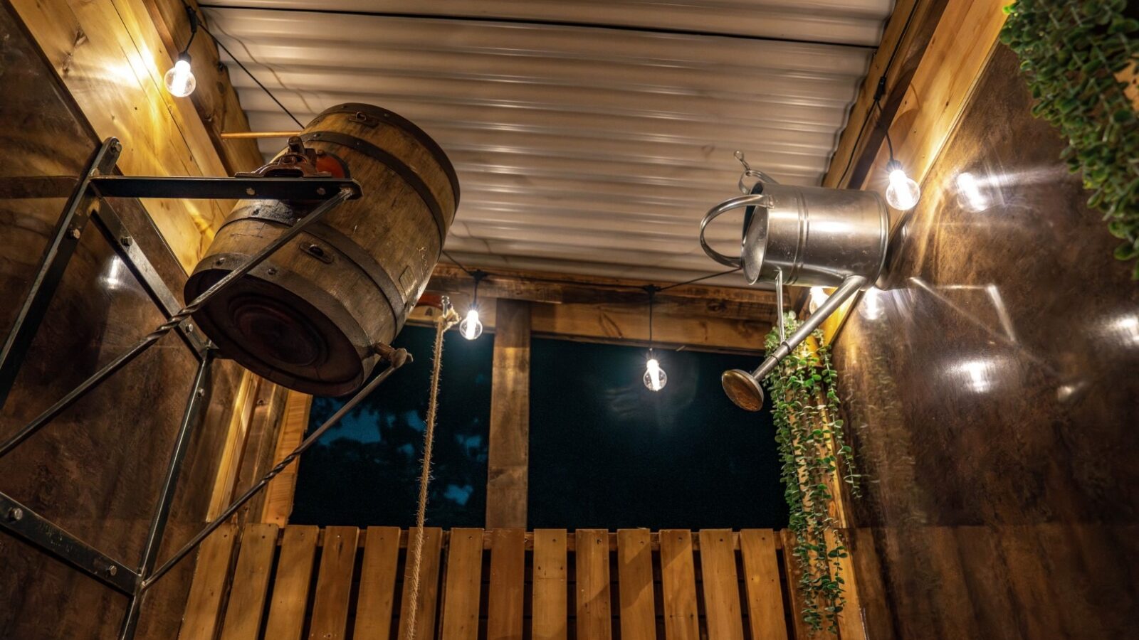 Discover the charm of Woodland Chase Cabins in this cozy wooden outdoor room, featuring a rustic barrel, watering can, string lights, and hanging vines that adorn its brown walls and corrugated metal roof.