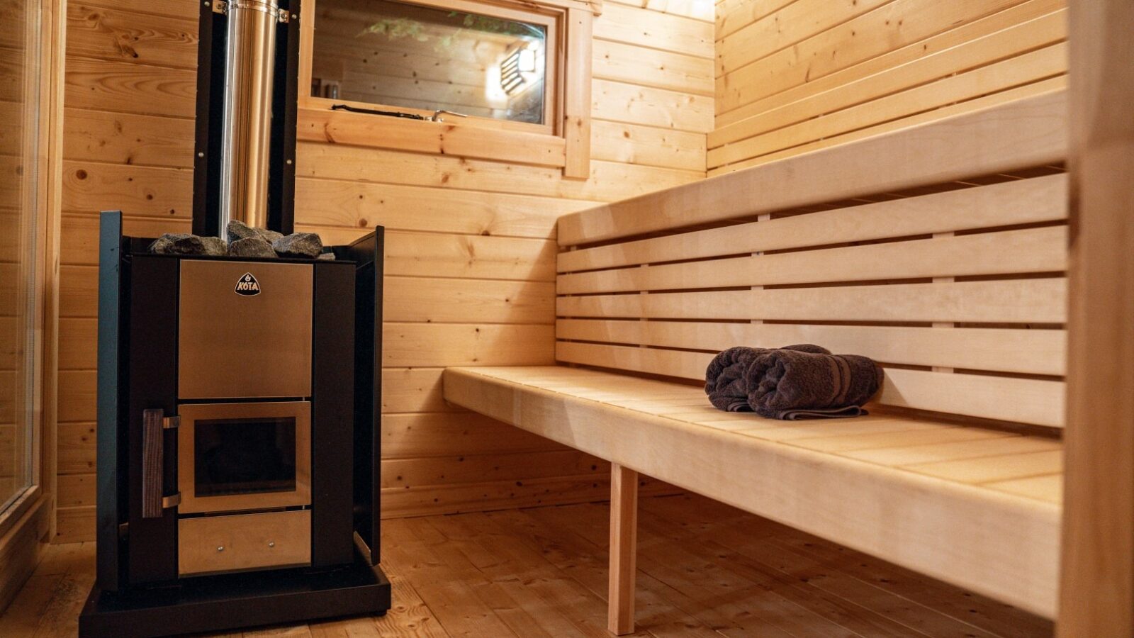 The Wooden sauna interior at Woodland Chase Cabins features a cozy bench, a neatly rolled towel, and a sleek black metal stove with chimney, offering an authentic cabin experience.