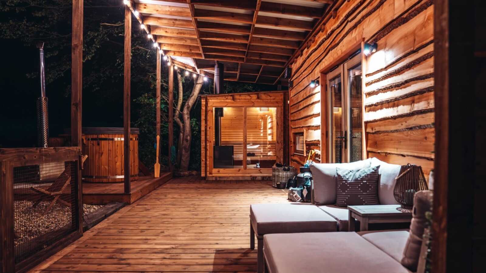 Beneath a canopy of stars, the wooden deck at Woodland Chase features a cozy sectional sofa adorned with twinkling string lights. The small cabin-like structure in the background adds to the enchanting atmosphere of this serene escape.