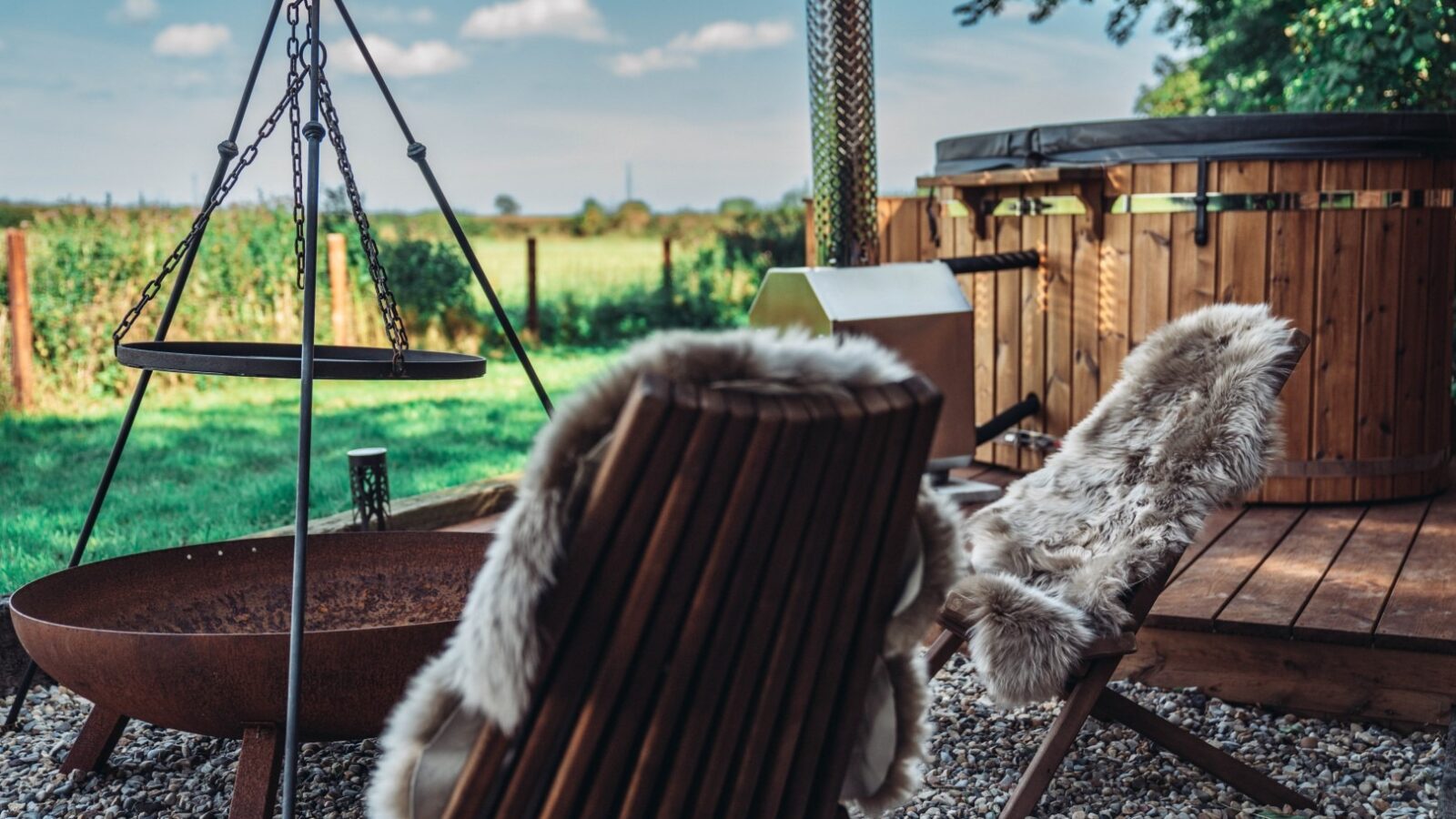 The outdoor seating area at Woodland Chase Cabins offers fur-covered chairs, a fire pit, and a hot tub, all with a scenic view of the grassy field and trees.