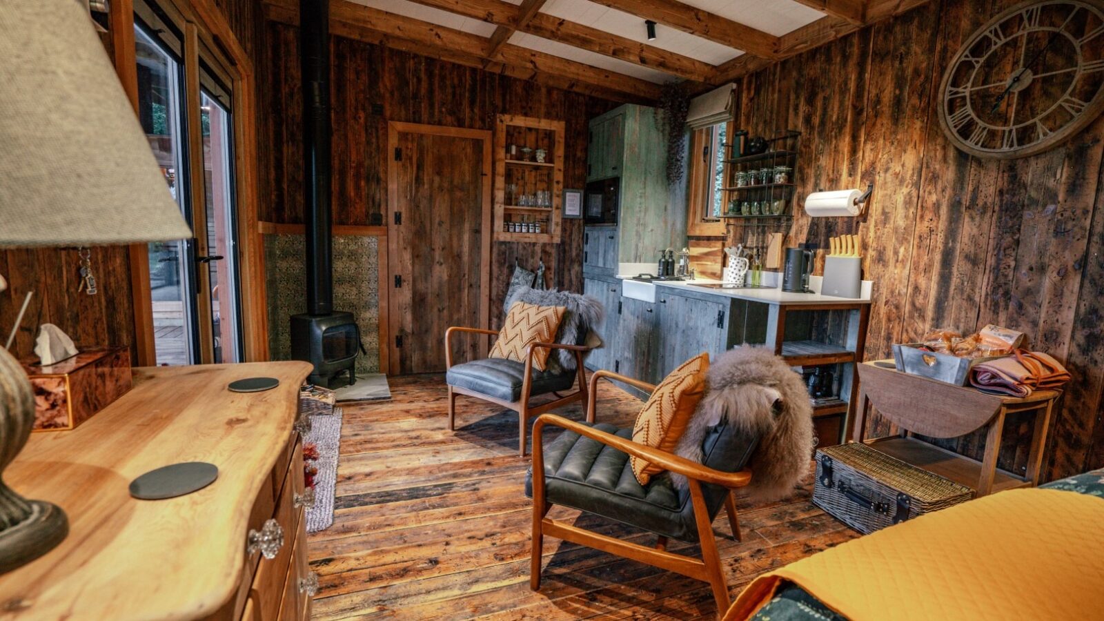 The cozy Woodland Chase cabin interior features a small kitchen, inviting chairs, a wood-burning stove, and a charming dresser, all under the watchful tick of a large wall clock.