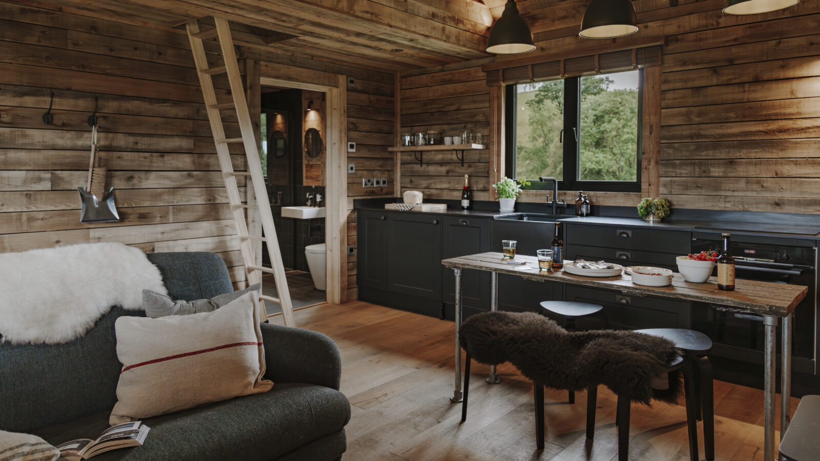 The cozy interior of Woolly Wood Cabins features wooden walls and floors. On the left, a green couch with a furry white throw invites relaxation, while on the right, a compact kitchen with black cabinets awaits. A small dining table with stools and pendant lights hangs from the ceiling near a partially open bathroom door.