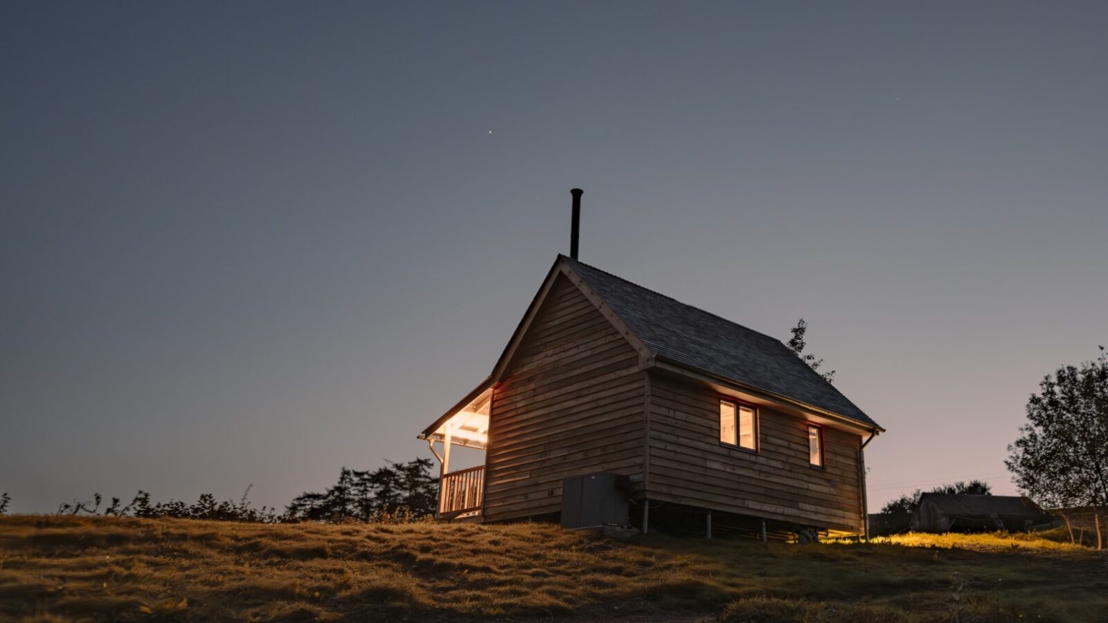 A small wooden cabin with warm lights glowing inside stands on a grassy hill under a clear night sky. The cozy cabin, adorned with woolly curtains, has a slanted roof and a chimney. A few trees are visible in the background, and the ground in the foreground is slightly illuminated.