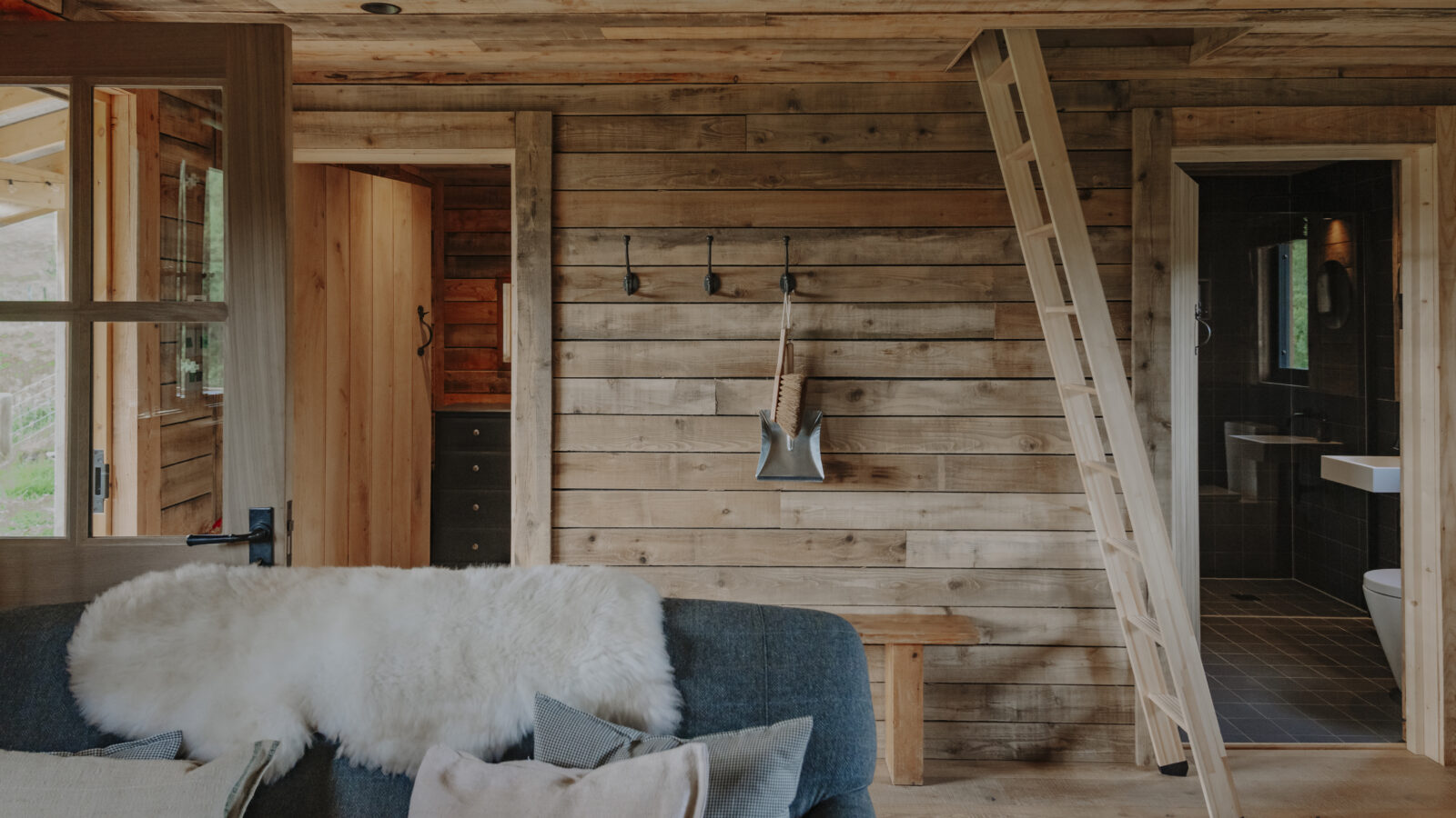 A cozy cabin interior with woolly touches, featuring wooden walls and ceiling. The room includes a ladder leading to a loft, a rustic hook rack with an axe hanging, a door open to a bathroom, and a blue couch topped with a fluffy white blanket and assorted pillows.