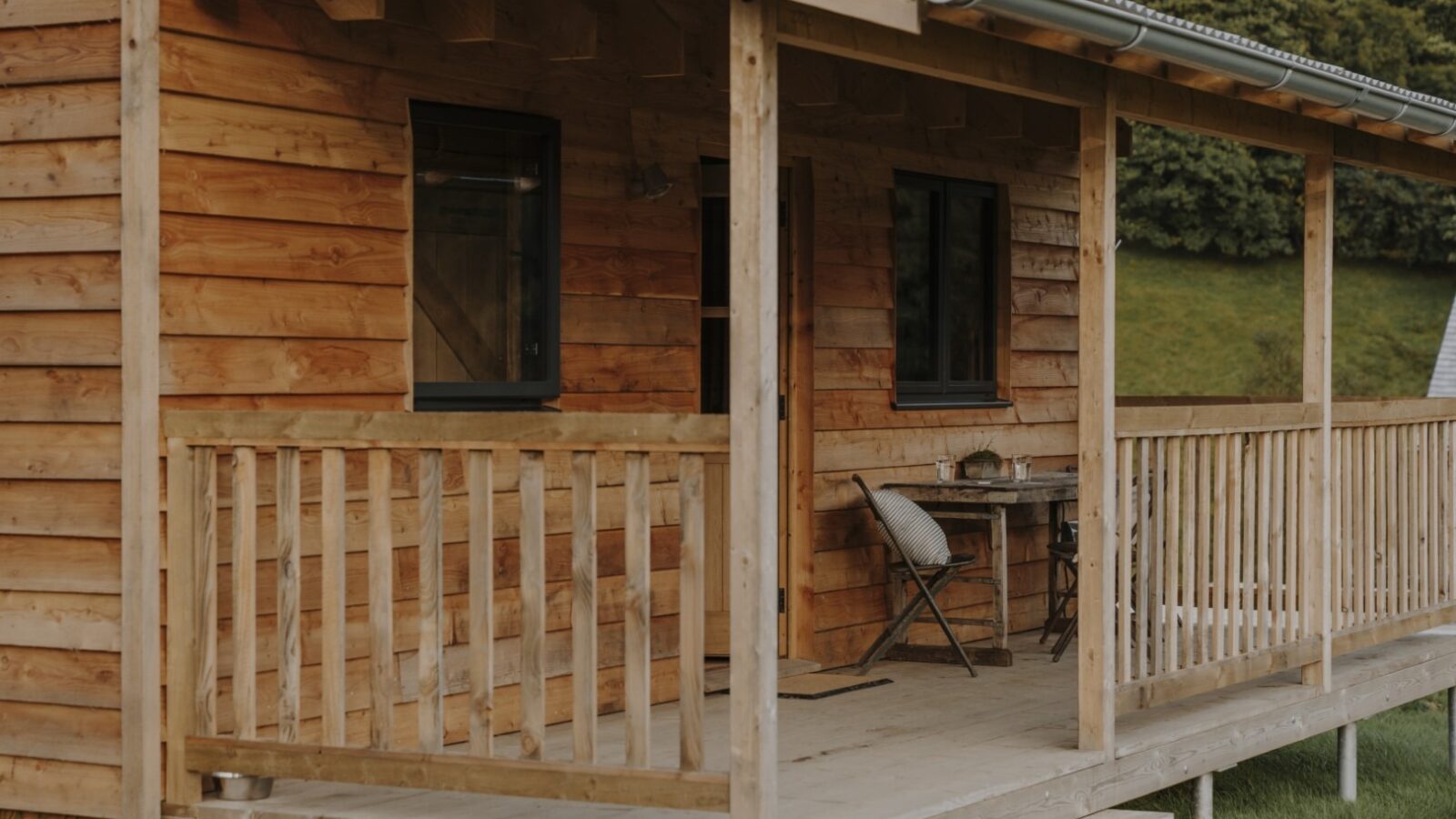 A rustic wooden cabin with a front porch featuring a table, a chair, and a woolly pillow. The porch railing, walls, and roof are made of natural wood. Trees and greenery are visible in the background, creating a serene, nature-filled setting perfect for those who love idyllic cabins.