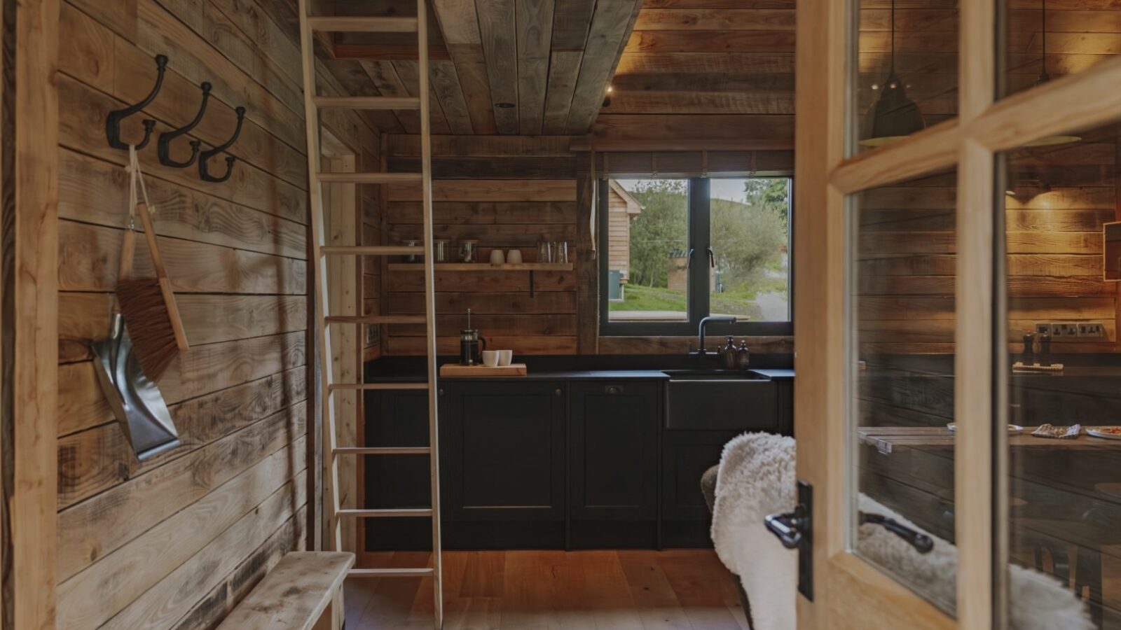 A cozy wooden cabin interior featuring a kitchen with a large window, black cabinets, and a sink. A wooden ladder leads to a loft area. Hooks on the wall hold various items, and a soft rug and woolly cushioned furniture add comfort. Natural light fills the space.