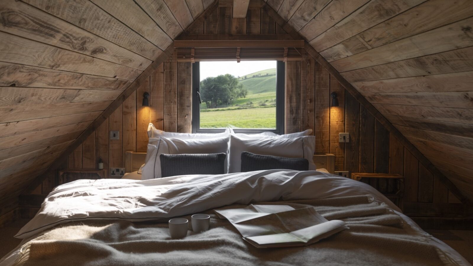 A cozy attic bedroom in the Woolly Wood Cabins features wooden walls and ceiling. A bed with rumpled white and beige linens faces a window that offers a view of green hills and trees. Two mugs and an open book rest on the bed, while warm lighting from bedside lamps creates an inviting atmosphere.