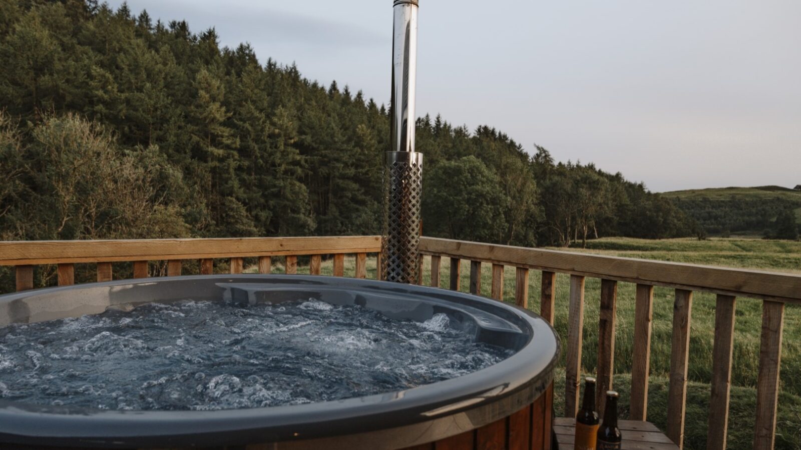 A bubbling hot tub is placed on a wooden deck overlooking a scenic landscape with dense green trees and open fields. Two beer bottles sit on the edge of the wood railing. The sky is clear with subtle hints of sunset or sunrise light, adding to the cozy vibe reminiscent of rustic cabins.