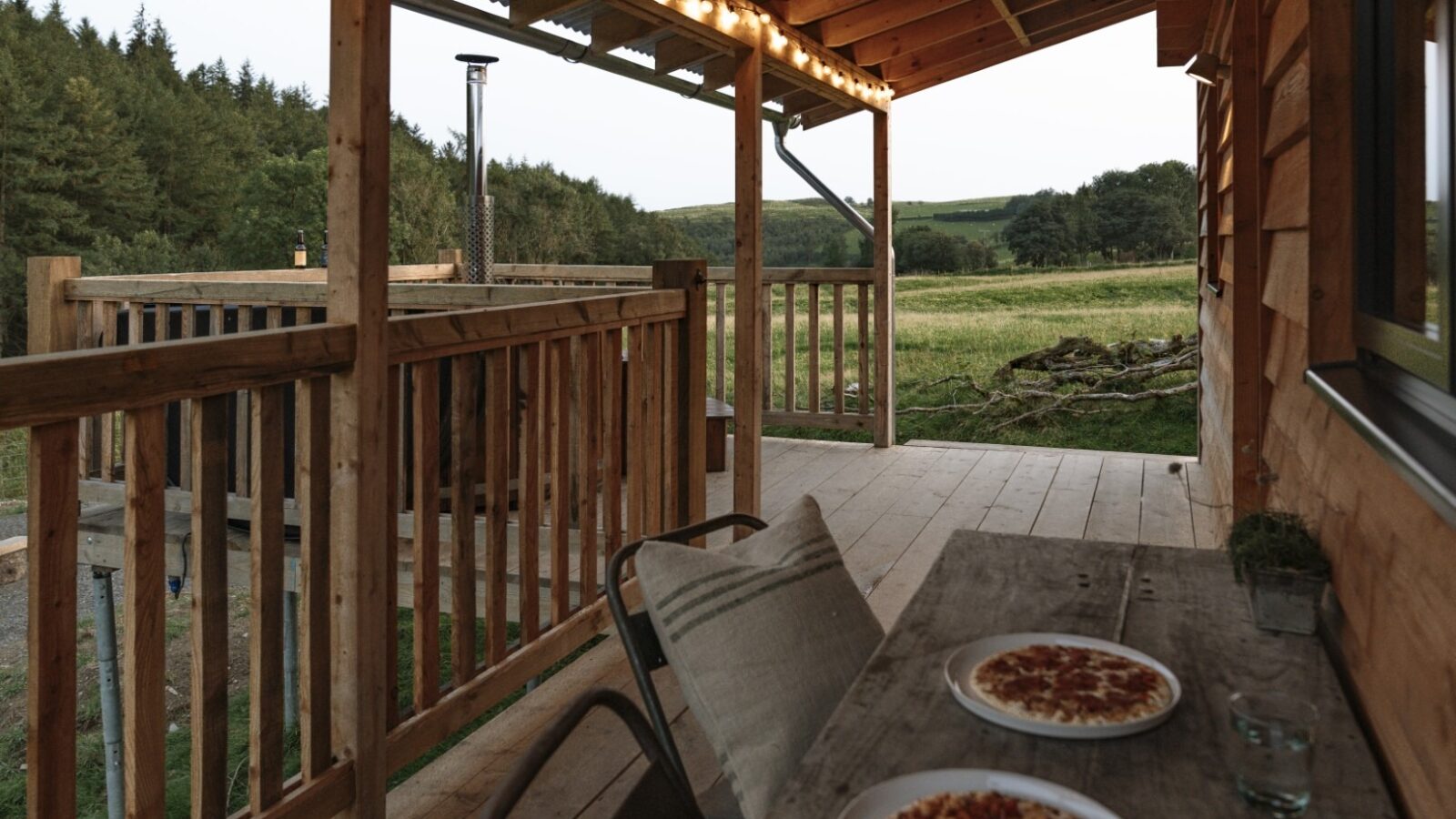 A wooden porch at Woolly Wood Cabins overlooks a serene rural landscape with green fields and trees. Two plates of pizza rest on a table with two chairs, and warm string lights are strung under the porch roof. The setting is calm and inviting, perfect for a relaxed outdoor meal.