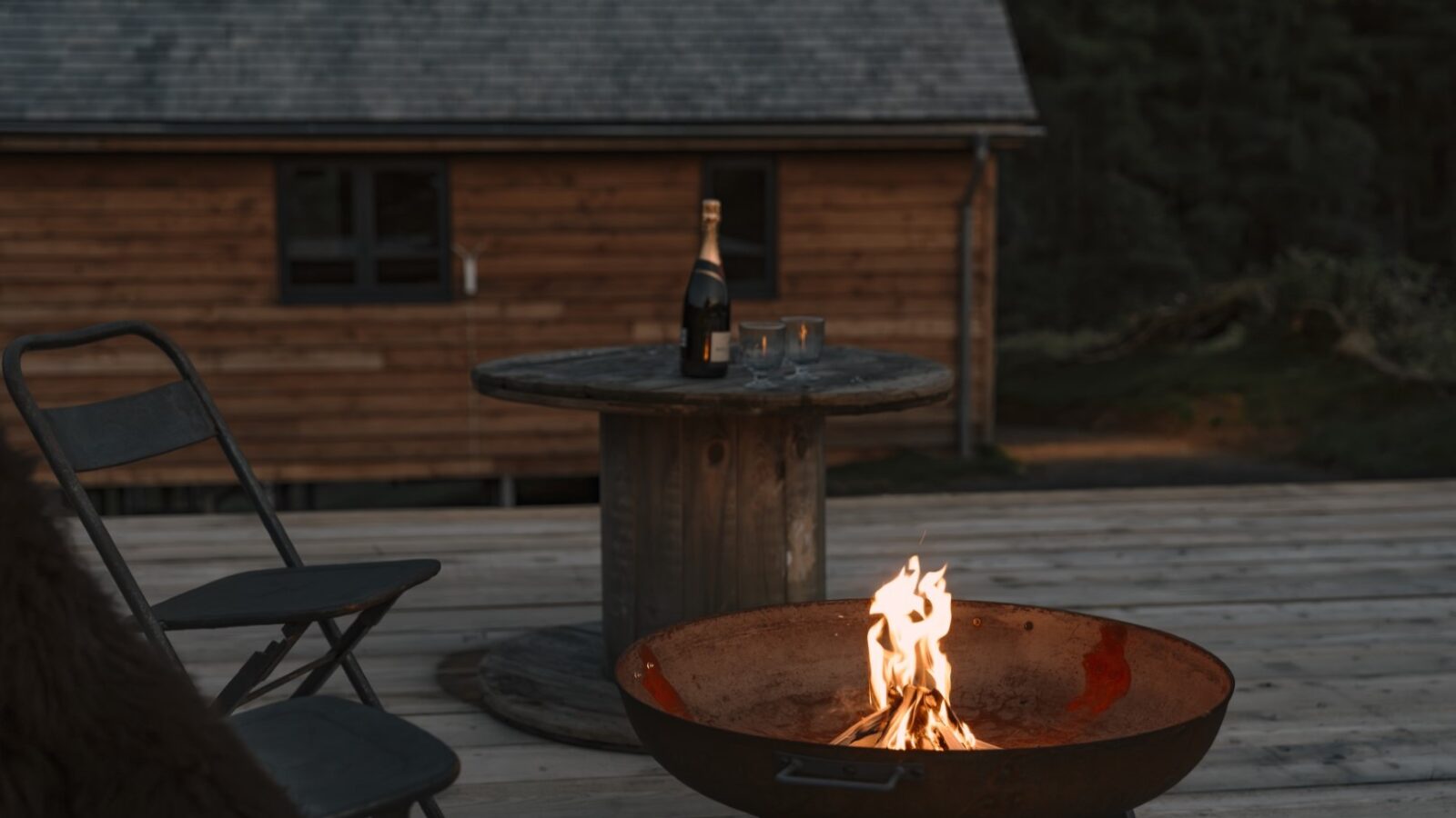 A cozy outdoor setting featuring a fire pit with a gentle flame, wooden chairs, and a spool table with a bottle of champagne and two filled glasses. The charming backdrop of Woolly Wood Cabins includes a shingled roofed cabin and lush forest beyond.