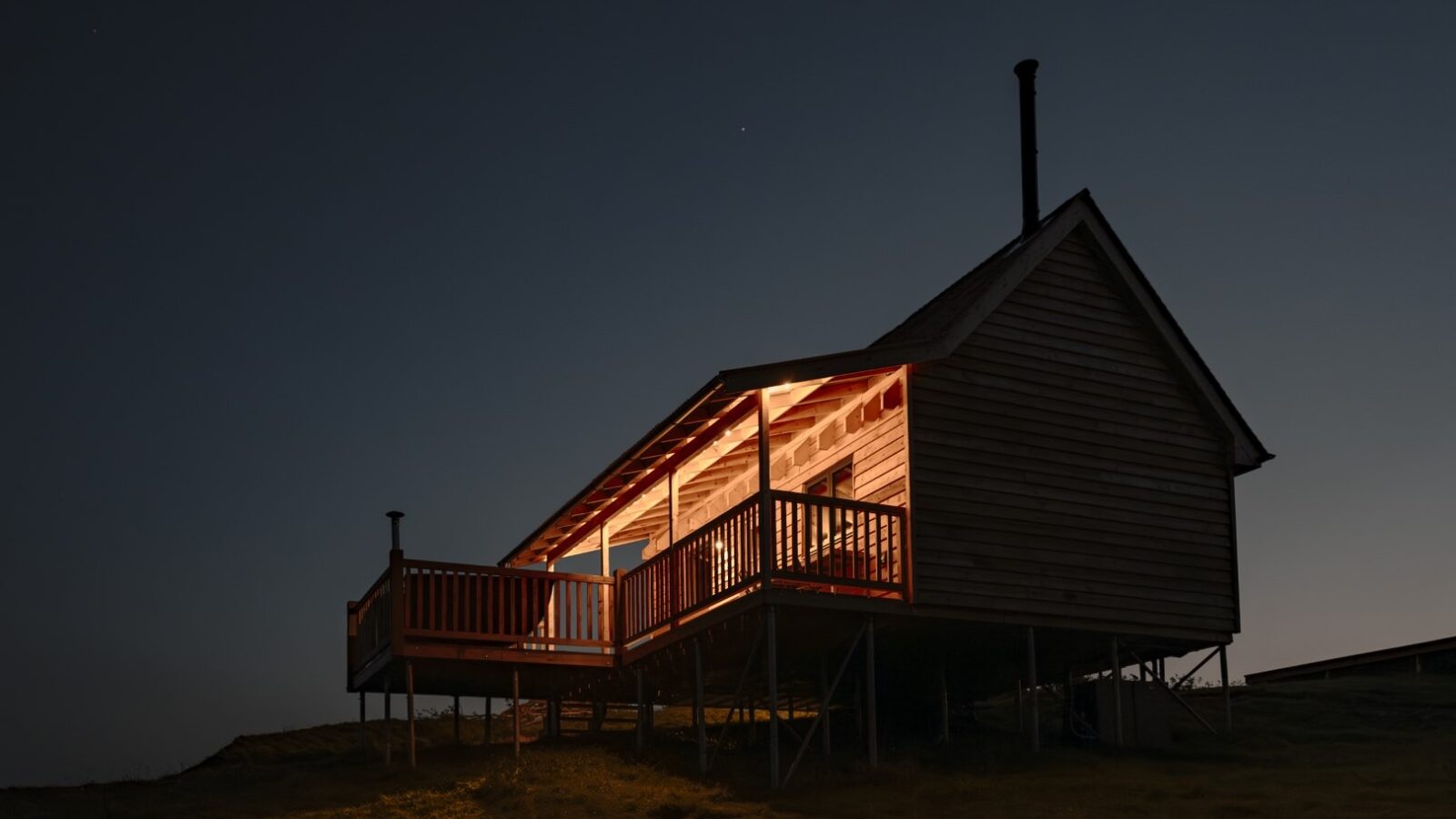 A small wooden cabin with a porch, part of the Woolly Wood Cabins collection, is warmly illuminated at night. The house is raised on stilts and stands alone in a dark, open area. The dark sky in the background is clear with a faint star visible.