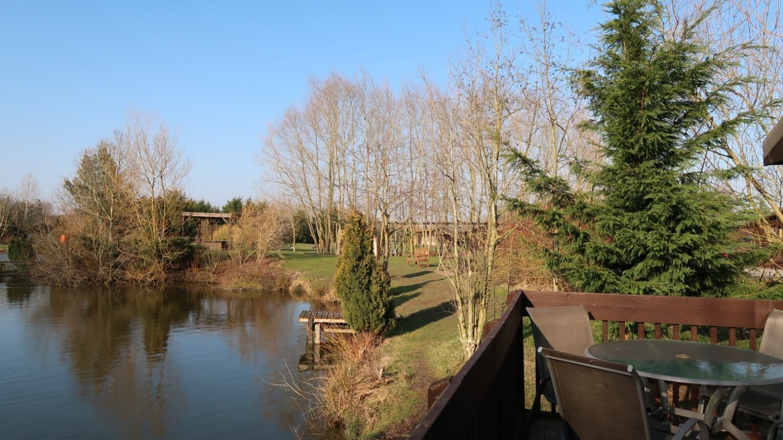 A scenic view of a calm pond in the Country Park, surrounded by bare trees under a clear blue sky. A wooden deck with a glass table and chairs overlooks the water. Sparse greenery and Yellowtop add a touch of color to the serene winter landscape.