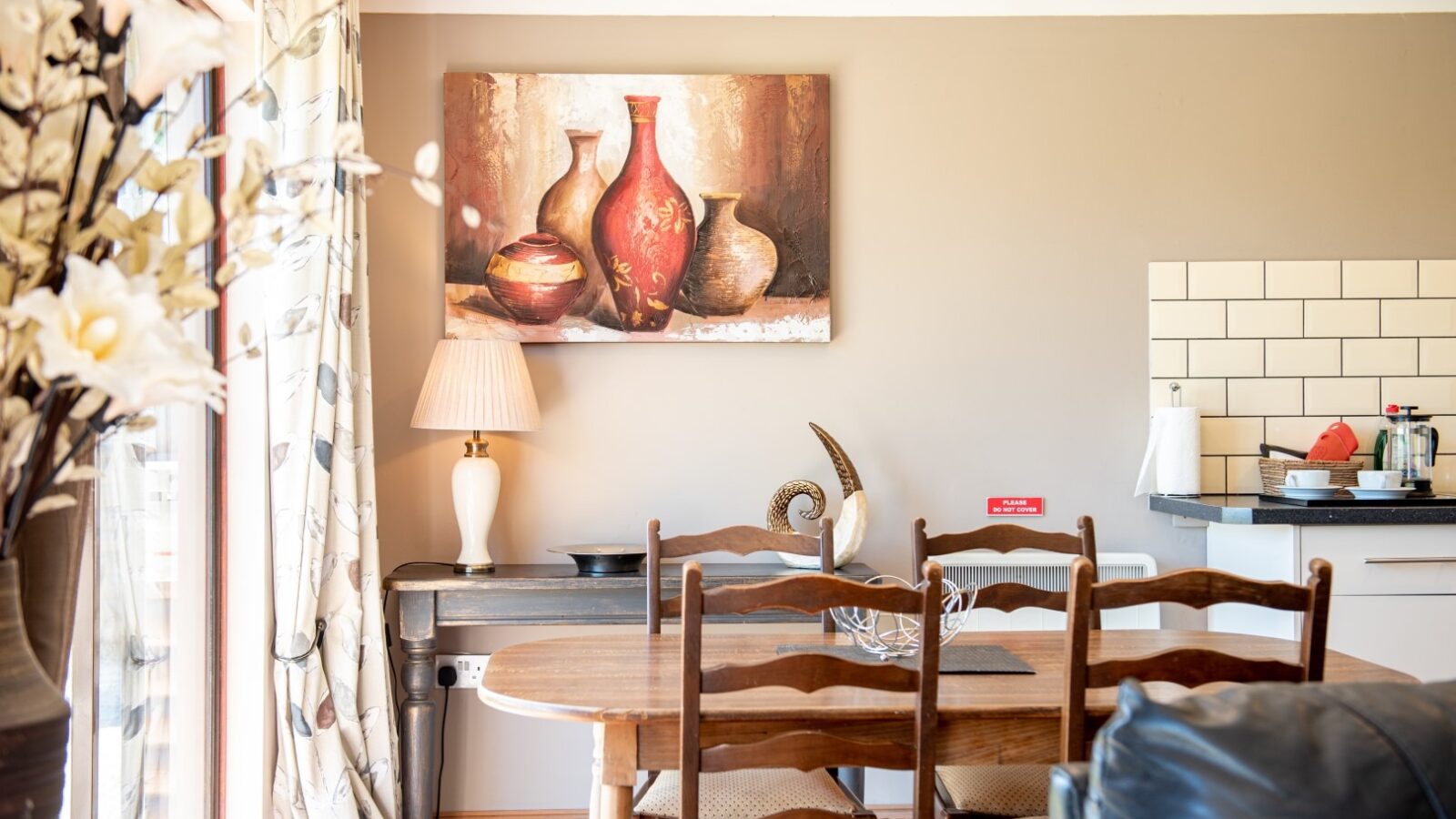 A cozy dining area with wooden chairs around a table. A painting of vases hangs on a beige wall above a wooden console table with a lamp and decorative items, reminiscent of Yellowtop. Nearby, a kitchen counter with a kettle and coffee maker is visible. Light from a window illuminates the space.