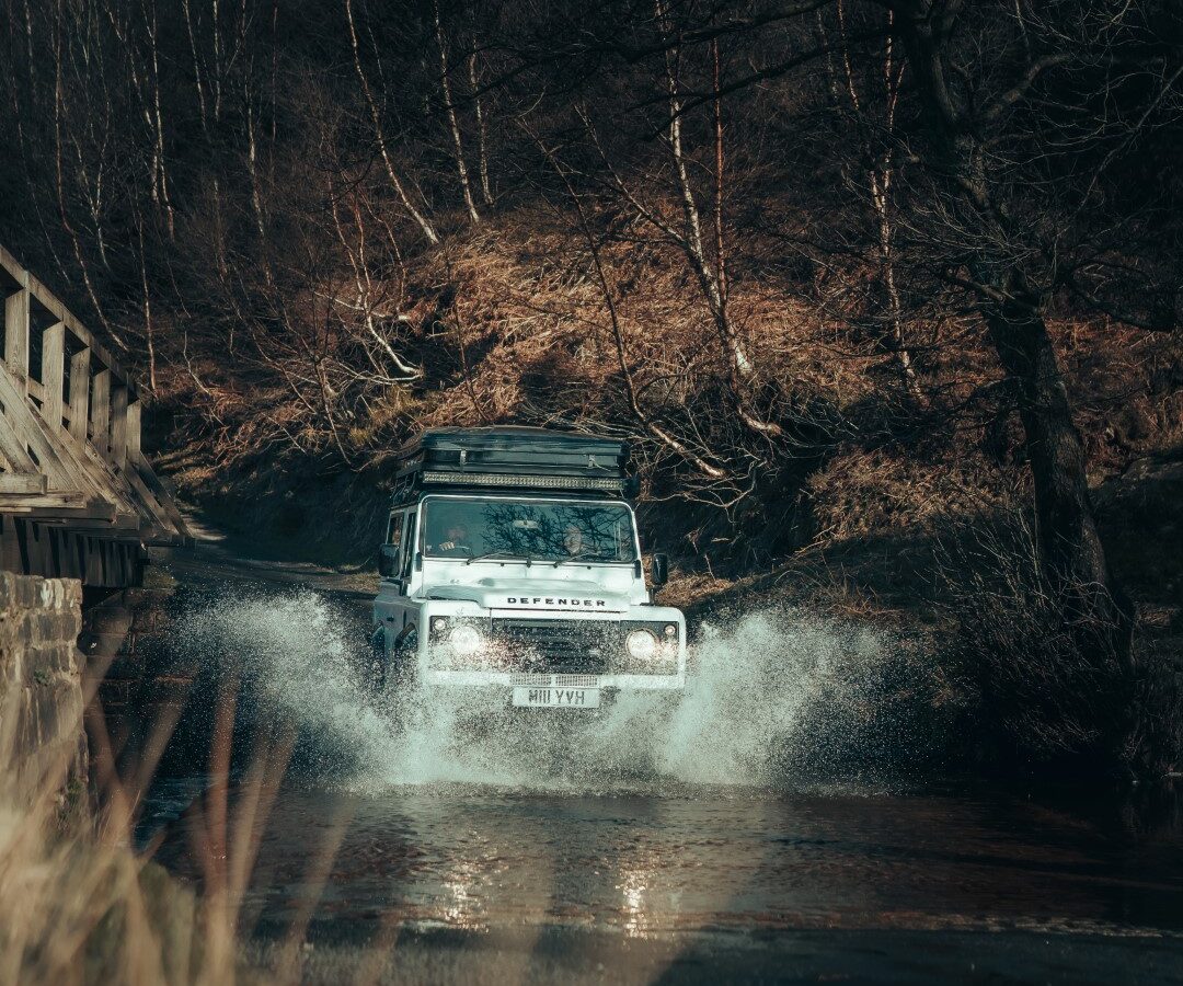 A white Defender SUV maneuvers through a shallow stream near a wooden bridge, with leafless trees silently surrounding the scene, hinting at an adventurous camping trip ahead.