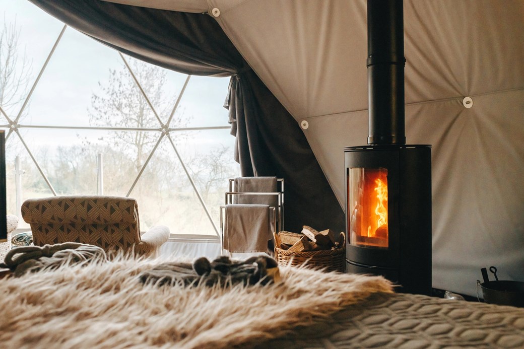 Inside the serene woodland escape, the geodesic dome features a cozy burning stove surrounded by chairs, a patterned armchair, and a plush fur throw near the triangular window.