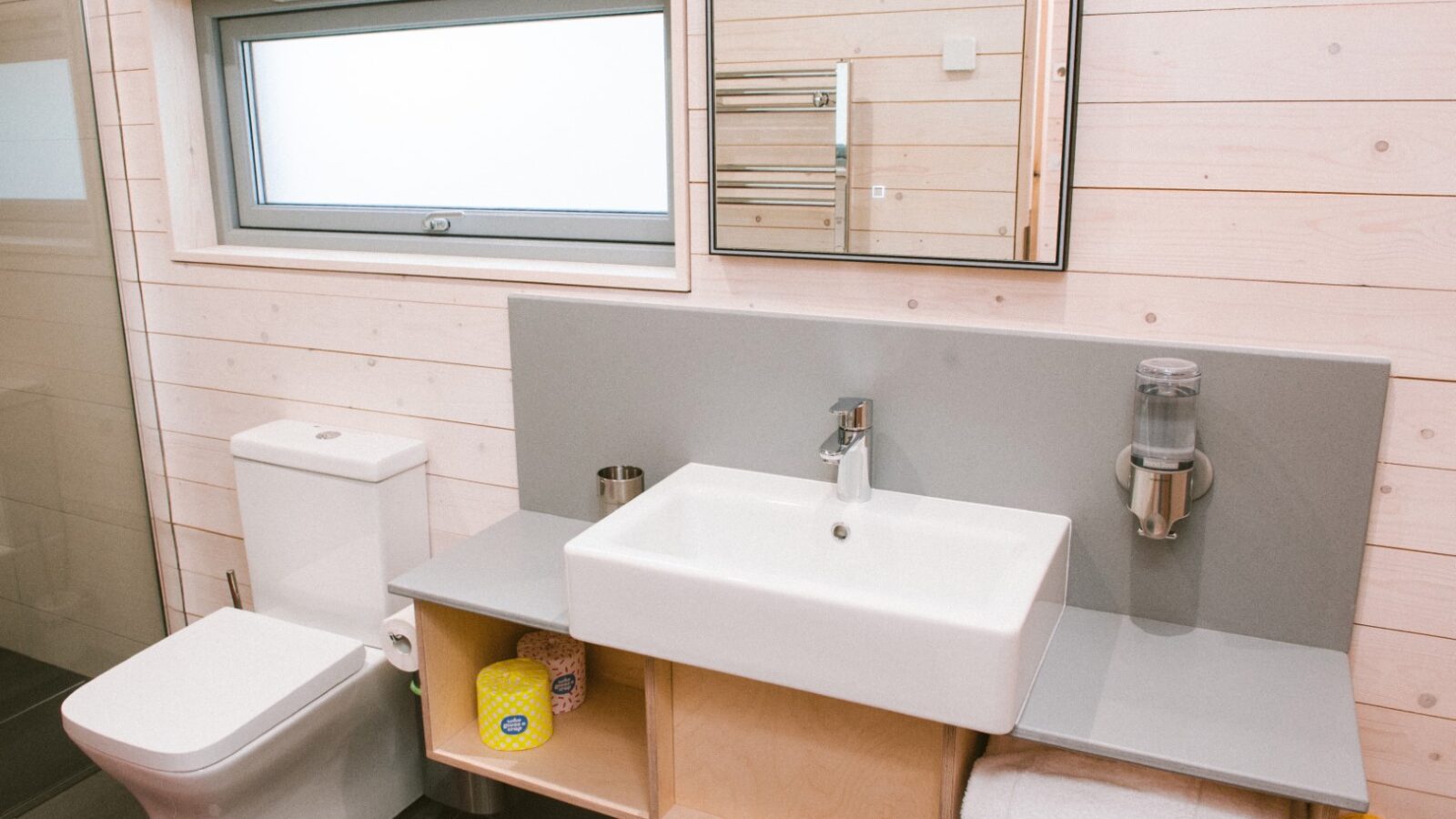 The Ecotone Cabins bathroom boasts modern design with light wood-paneled walls. It features a rectangular sink beneath a sleek mirror, a toilet, and a compact shelf for toiletries and towels. A frosted window ensures ample natural light.