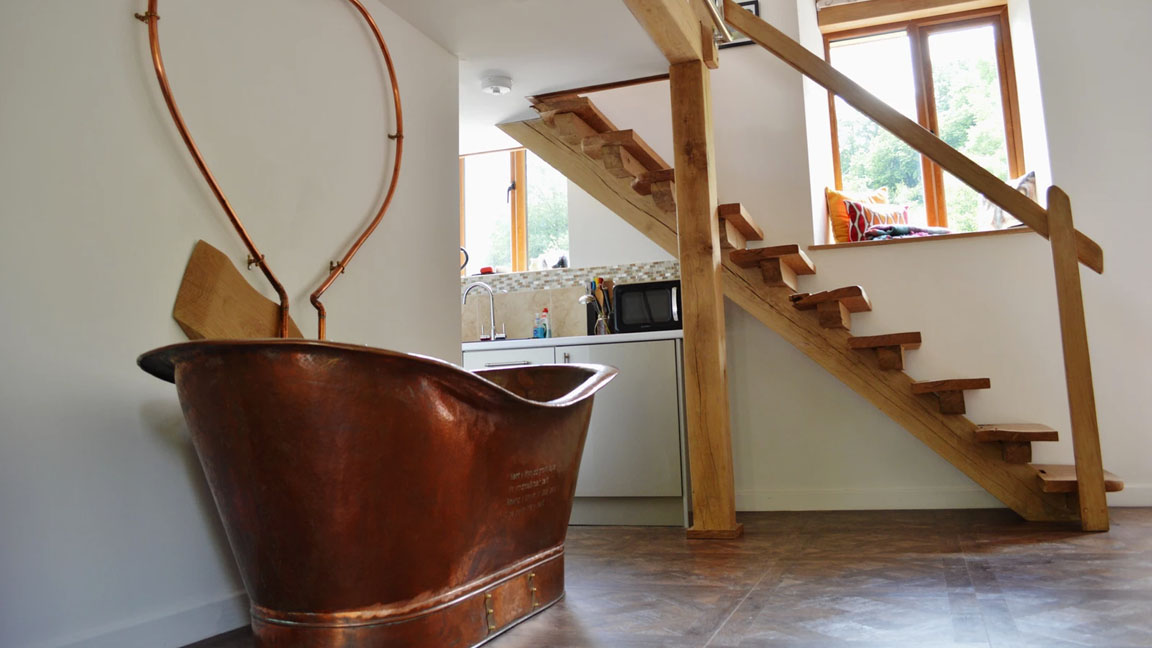 The elegant copper bathtub sits near a wooden staircase in the modern room of Nant Awen Cottages, accompanied by a quaint kitchen area and a sunlit window.