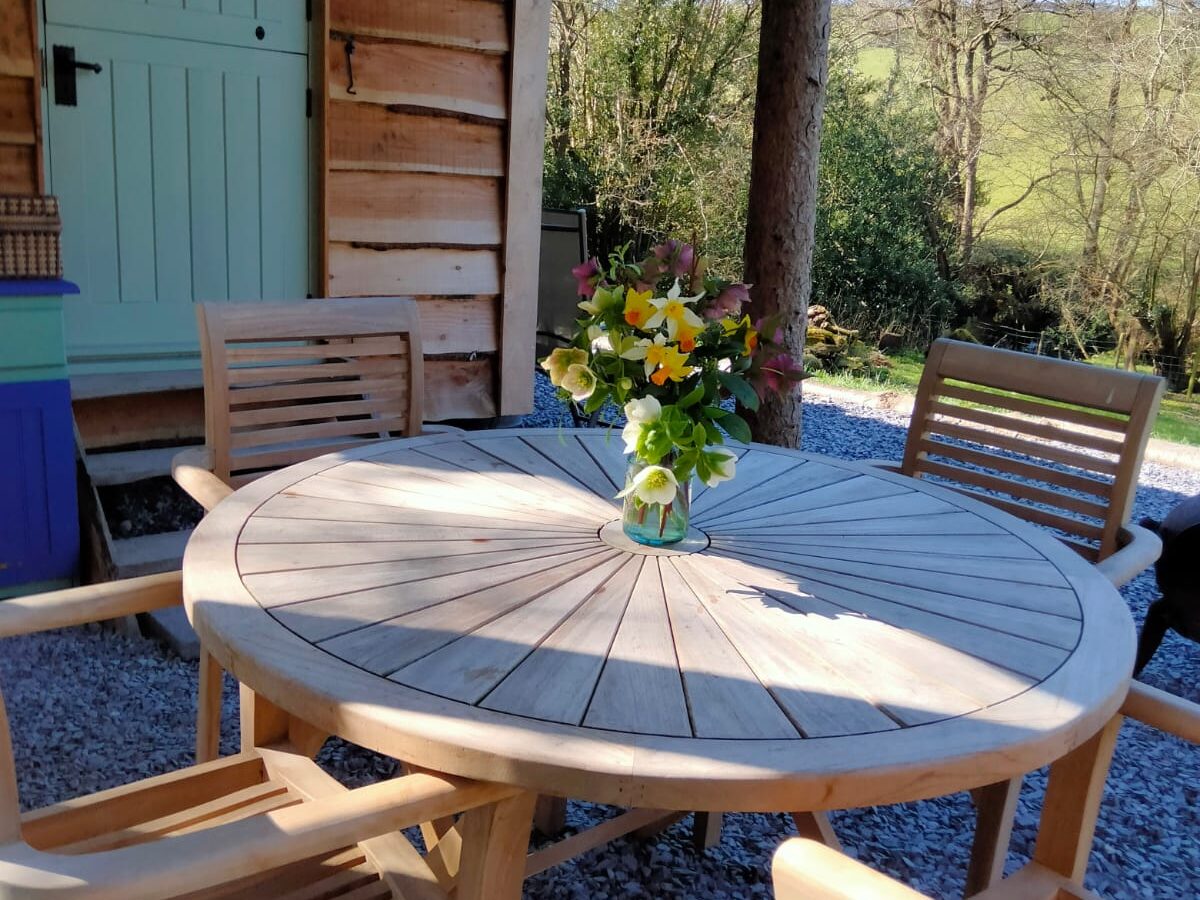 A wooden table with a vase of flowers, surrounded by chairs on a gravel patio, exudes rustic charm. The serene setting is completed by the cozy Caban Eric nestled among the trees in the background.