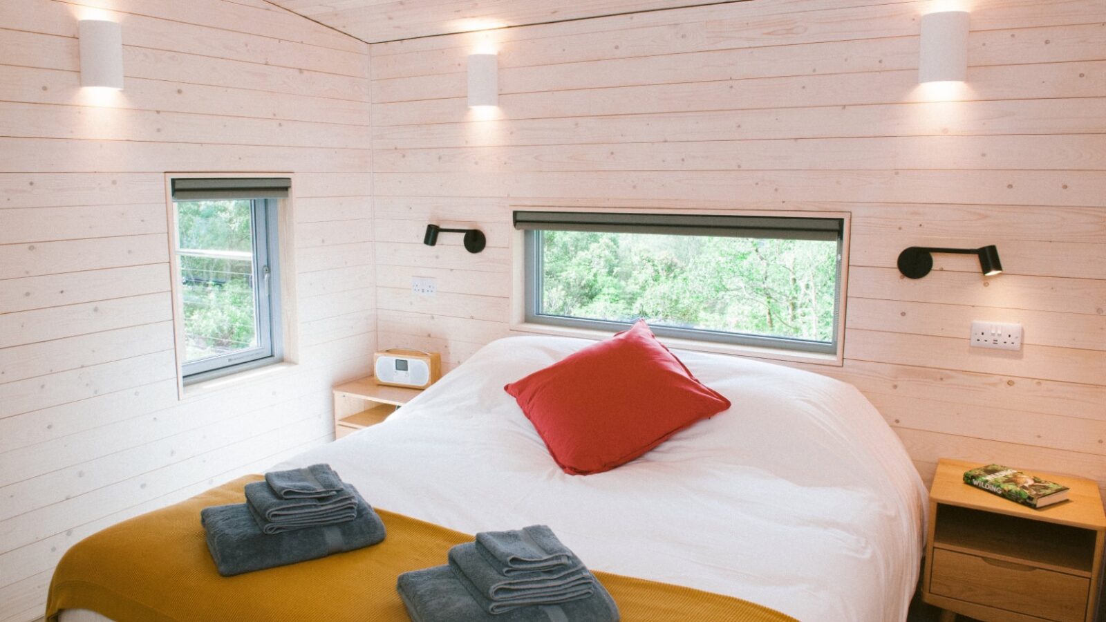 A cozy eco-friendly cabin bedroom with light wooden walls, featuring a large bed adorned with a red pillow and mustard blanket. Two neatly folded towels sit at the foot of the bed. Natural light streams through two windows, and small bedside tables with lamps complete this sustainable retreat.