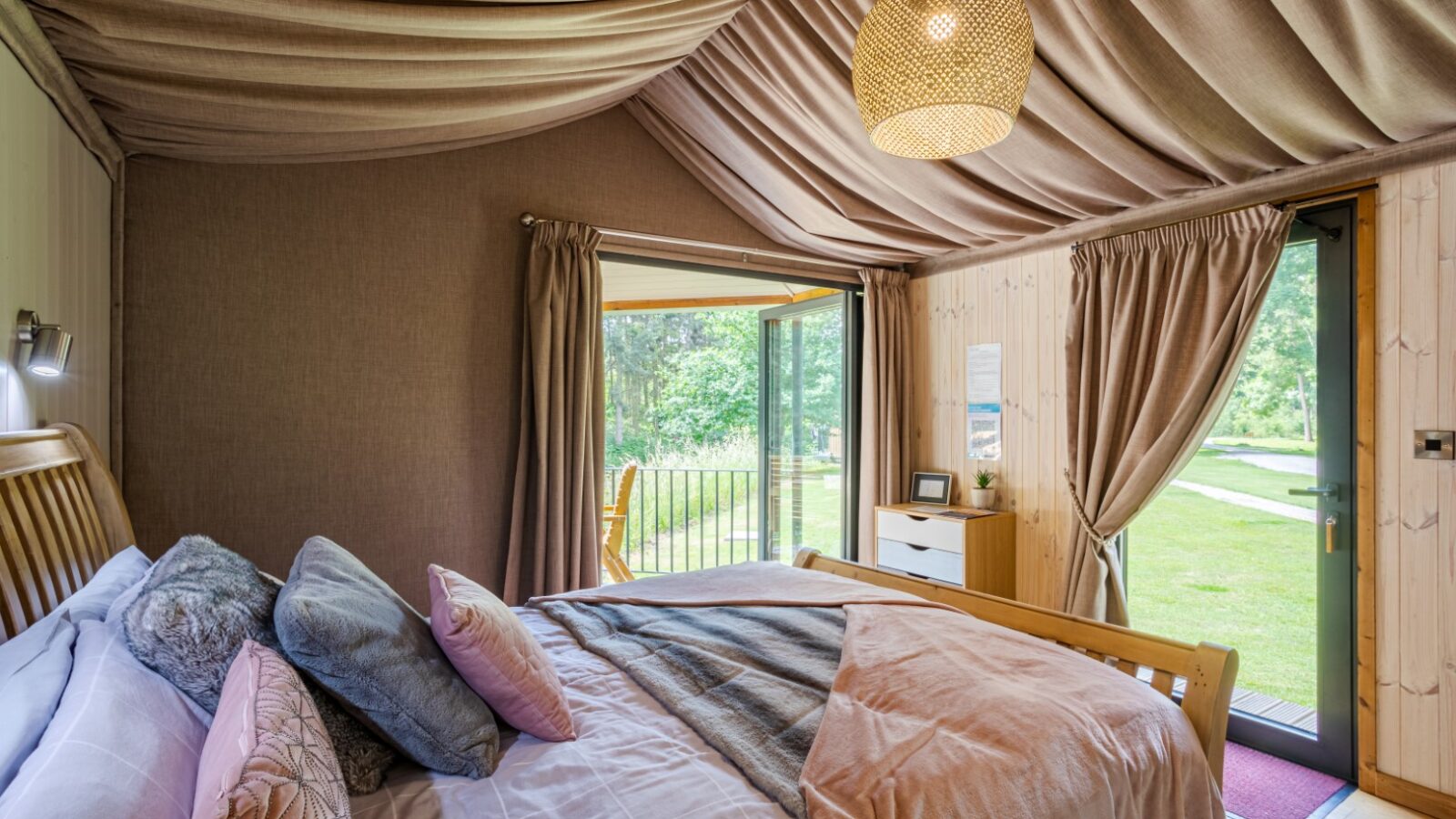 A cozy riverside bedroom with a draped ceiling, wooden bed, large windows, and doors opening to a garden view. The neutral-toned decor complements the serene surroundings, offering a tranquil retreat from the everyday hustle and bustle.