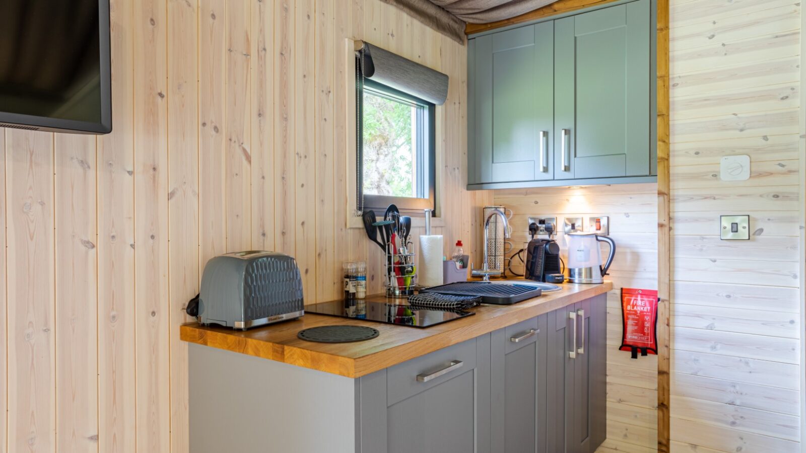 This compact riverside kitchen boasts gray cabinets, a toaster, two-burner cooktop, kettle, and utensils neatly arranged on a wooden countertop. Perfect for cozy cabins.