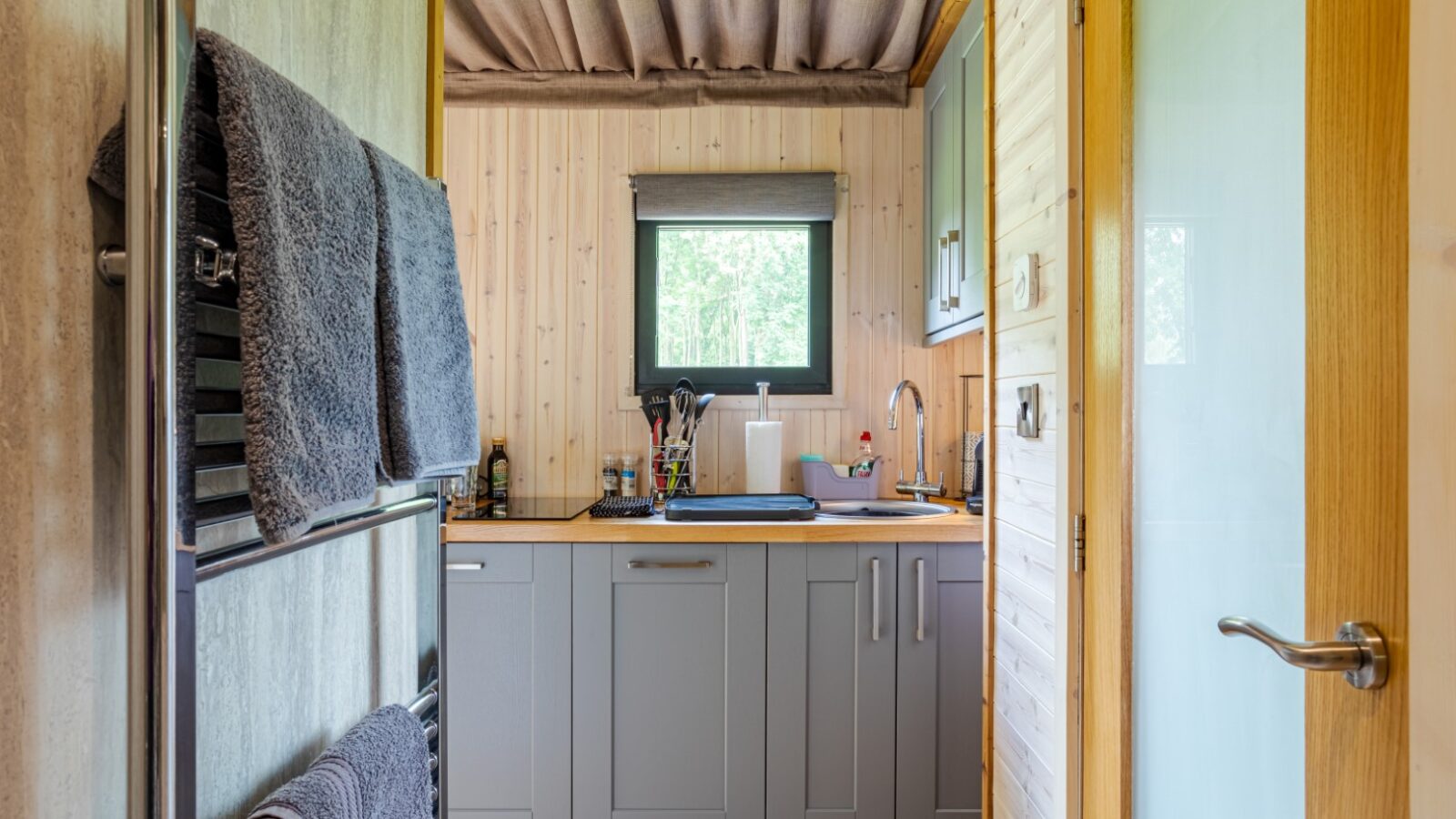 View of a compact laundry room with grey cabinets, reminiscent of cozy cabins, a countertop with household items, and towels on a rack by the door.