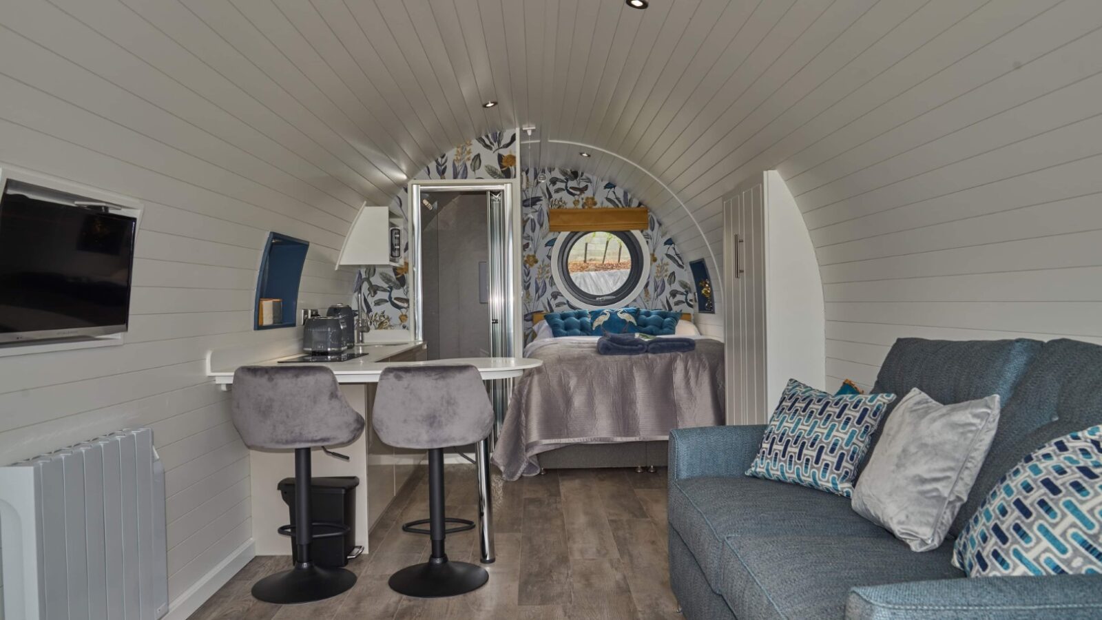 A cozy, modern tiny house pod with curved white walls offers a touch of luxury. It features a small kitchenette with two stools, a blue sofa adorned with patterned pillows, and a neatly made bed at the back. Natural light from Camlad Valley streams in through a small round window.