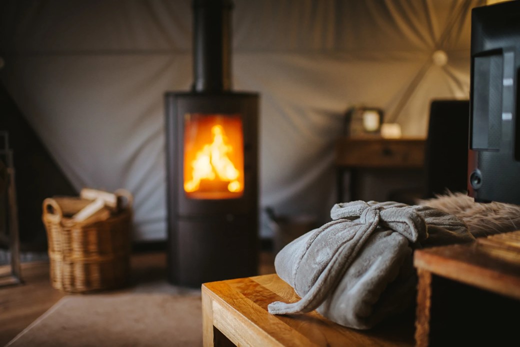 A cozy woodland escape features a lit wood-burning stove, a basket of logs, and a soft blanket draped over a wooden table in the foreground, perfect for those seeking tranquility in charming domes.