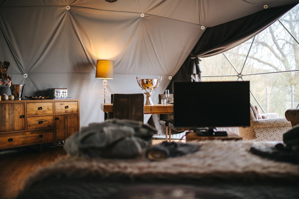 Cozy living space in a geodesic dome, perfect for Woodland Escapes, featuring a wooden dresser, lamp, dining table, and TV. A large window reveals stunning outdoor scenery.