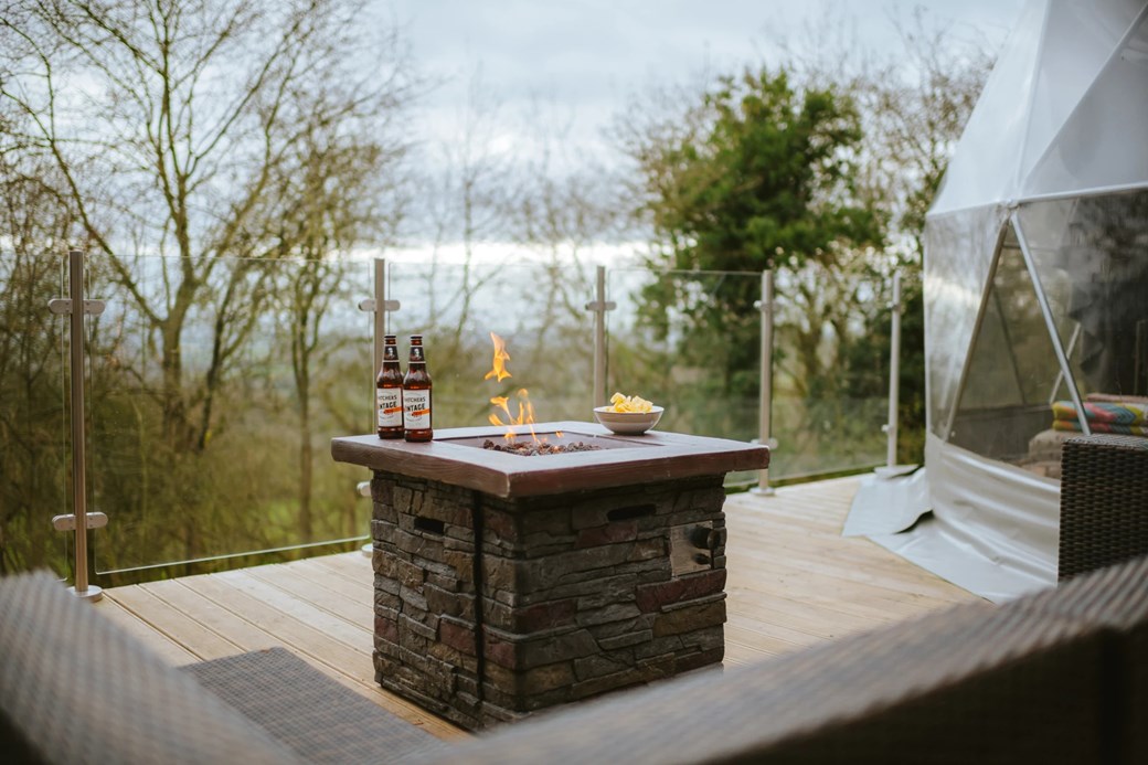 A patio with a stone fire pit, two beer bottles, and a bowl sits amidst the serene Woodland Escapes. Trees surround the area, with a tent-like structure resembling charming domes in the background.