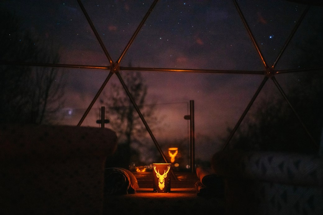 A candle glows inside a woodland dome with a starry night sky in the background, creating a cozy escape.