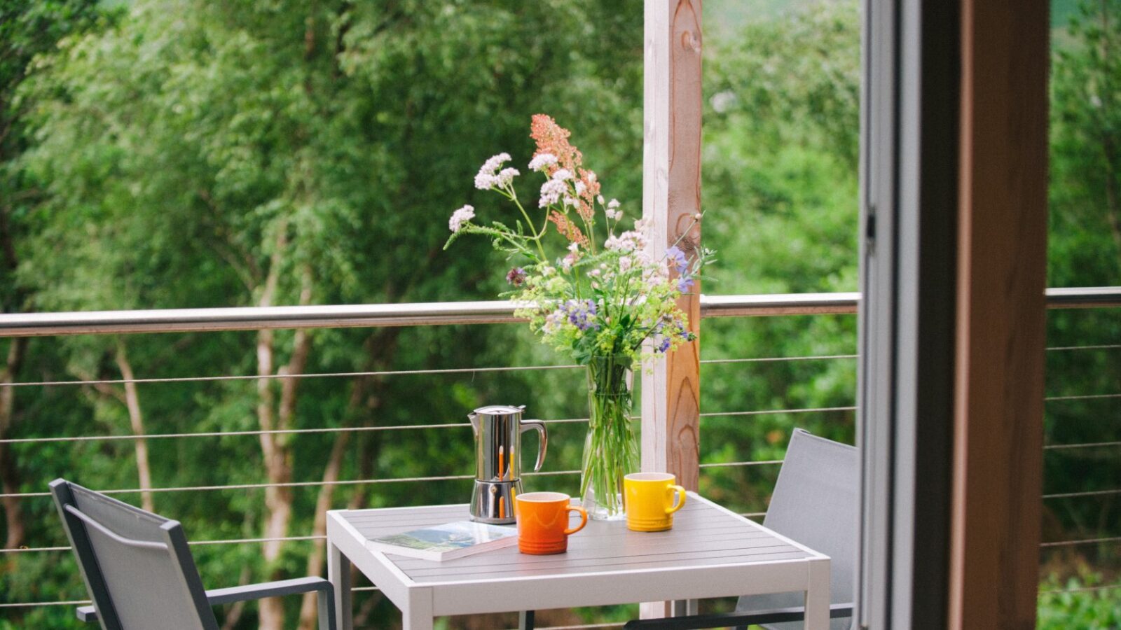 A small outdoor table on a balcony at Ecotone Cabins holds a bouquet of wildflowers in a vase, a silver coffee pot, an open book, and two colorful mugs. Surrounding the balcony, lush green trees provide a serene and natural backdrop for this eco-friendly nature retreat.