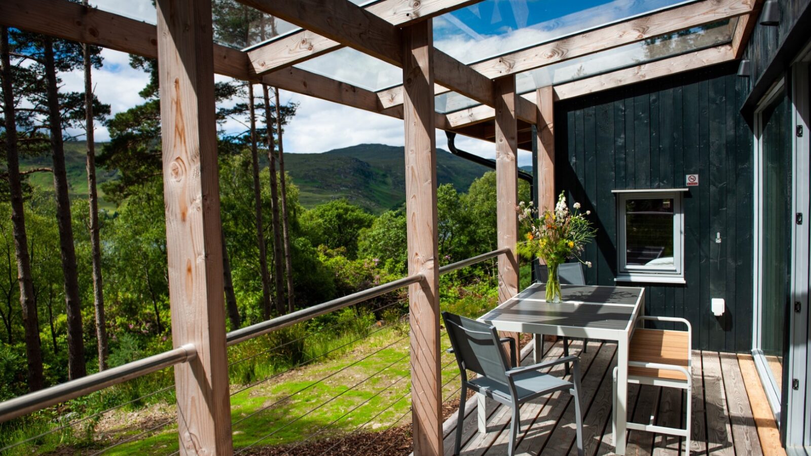 A cozy wooden deck with a table and two chairs sits under a clear roof, overlooking a lush ecotone of forest and distant hills. A vase with flowers adorns the table, and sunlight filters through the glass-paneled roof, enhancing the serene outdoor setting.