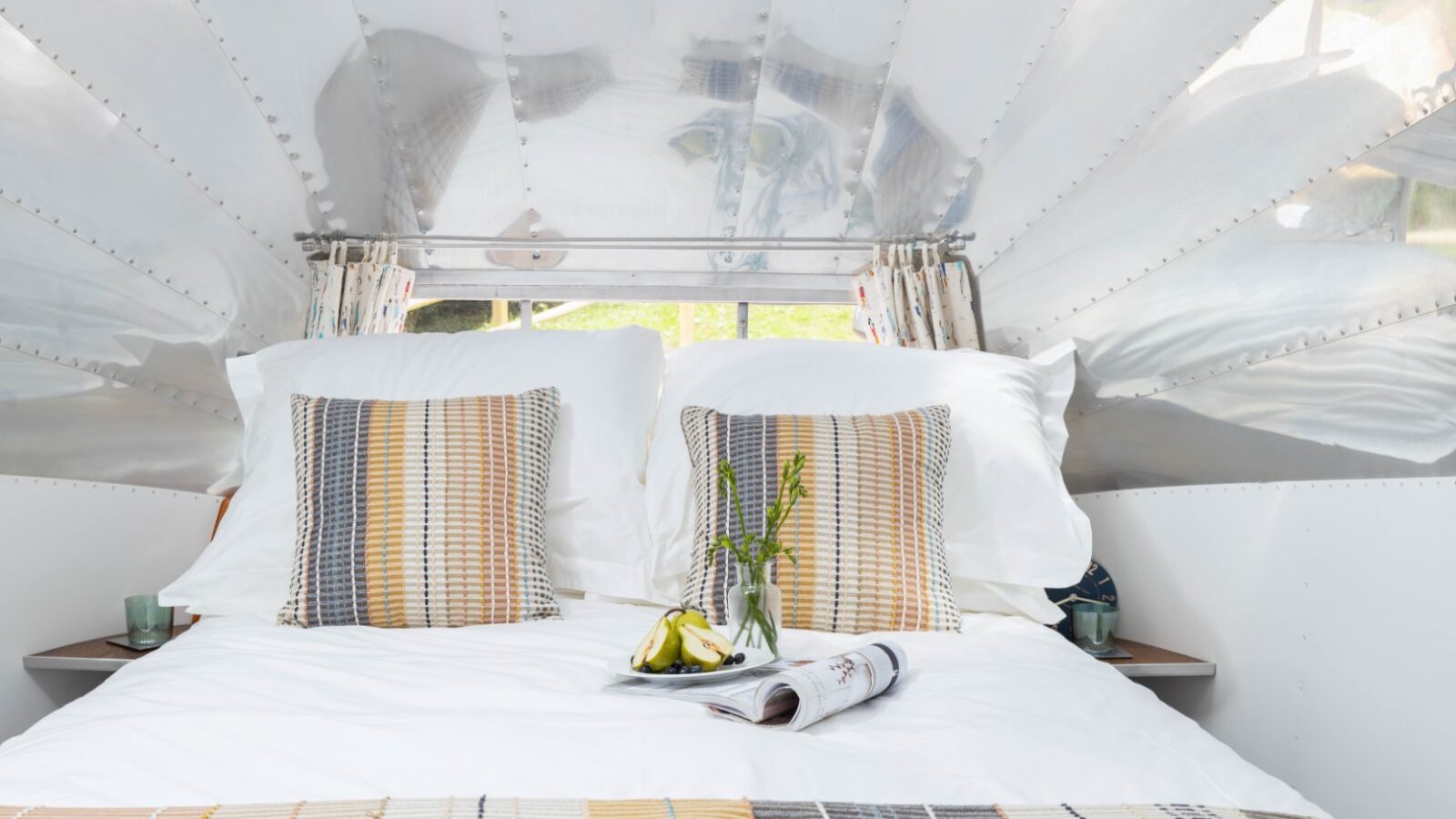 Inside a cozy, American-style metallic Airstream trailer, a bed with white sheets and pillows adorned with colorful striped accents awaits. A small vase of flowers, a magazine, and two pears rest on the bed as natural light streams through the windows.