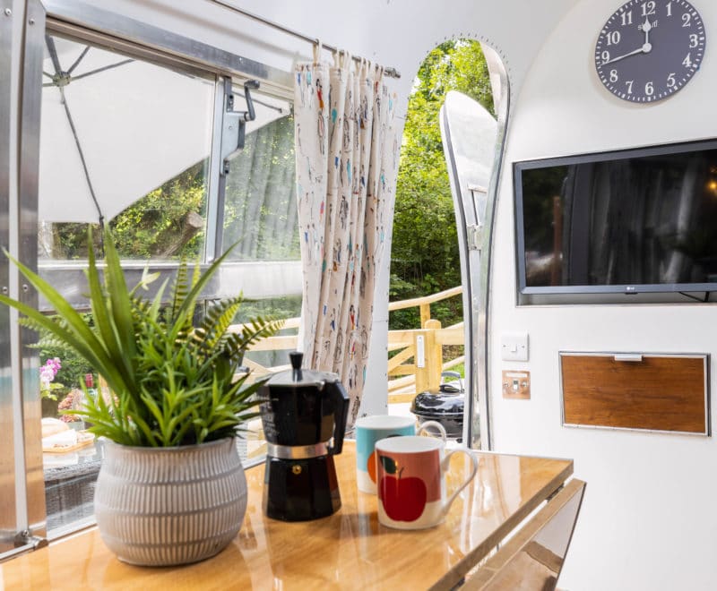 Inside a cozy Airstream, a window reveals lush greenery outside. A clock and TV are mounted on the wall. On the table sits a potted plant, a black coffee maker, and two mugs—one with an apple design. Light floral curtains frame the view, offering an inviting American charm to this Dittisham retreat.