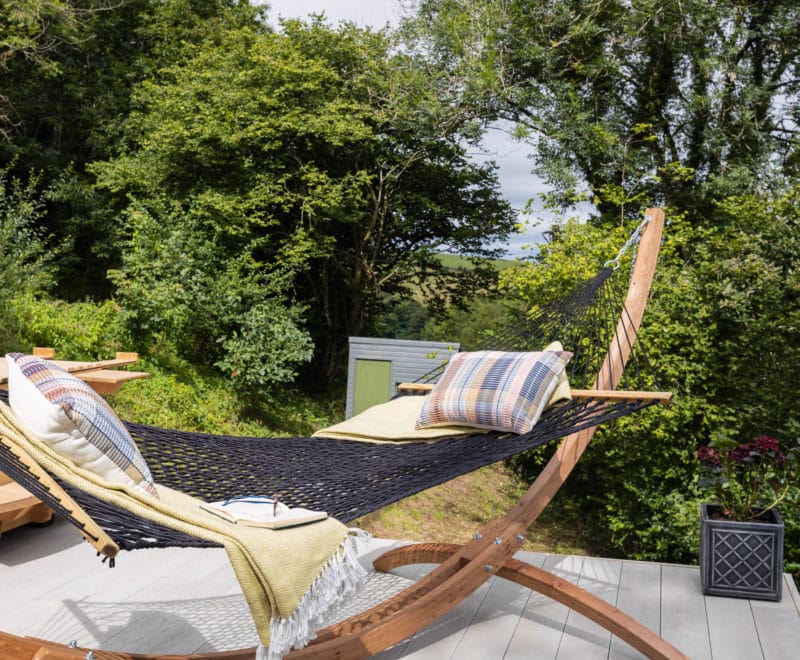 A cozy hammock with pillows and a blanket is set on a wooden stand on a patio. It overlooks lush greenery and an Airstream trailer in the background, creating a relaxing outdoor setting reminiscent of an idyllic American retreat.