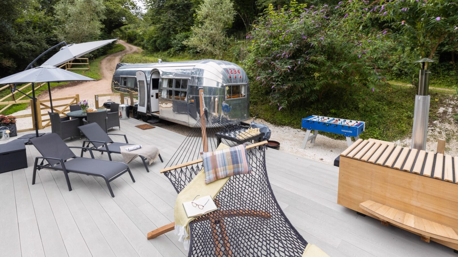 A cozy outdoor setting features a hammock, sun loungers, a foosball table, and an American Airstream trailer. The wooden deck has a small table with chairs and is surrounded by Dittisham's lush greenery and trees, creating a peaceful atmosphere.