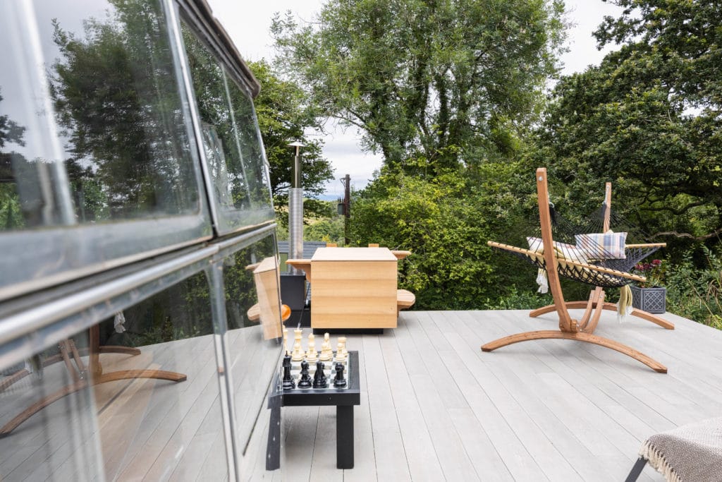 A scenic outdoor deck in Dittisham features a wooden dining table with chairs, an American black-and-white chess set, and a hammock strung between two wooden stands. Lush green trees surround the area, and the edge of a glass window is visible in the foreground.