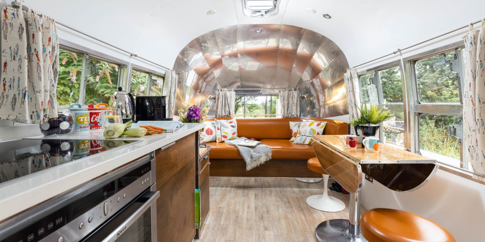 Inside a vintage Airstream trailer, the modern kitchen and living space exude an American charm. It features a brown leather couch adorned with colorful pillows, a small wooden table with chairs, and windows dressed in patterned curtains. Light wood flooring completes the stylish Dittisham-inspired look.