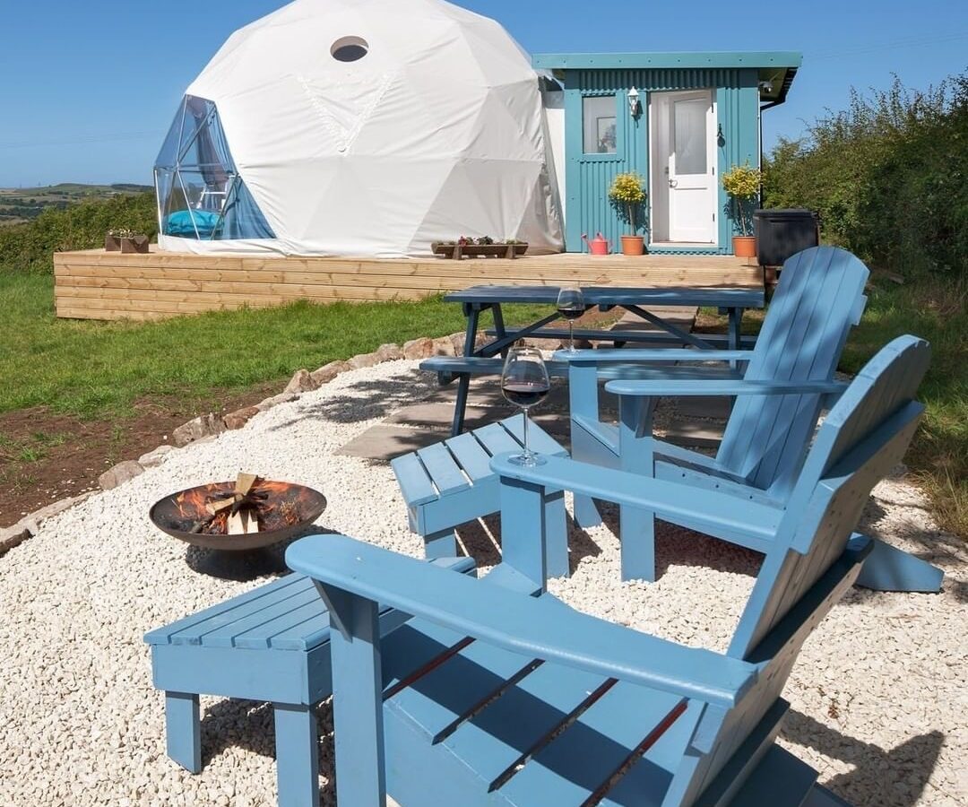 Blue Adirondack chairs surround a fire pit in front of the Mid Auchengowan Dome and a small blue cabin, all set on a grassy area under a clear sky.