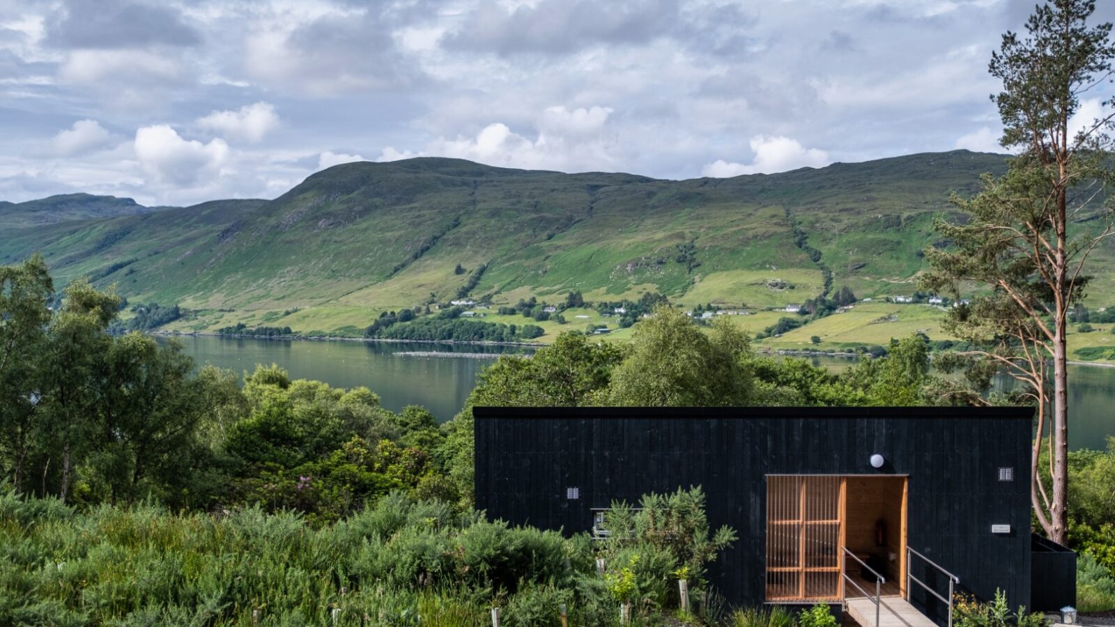 A modern black cabin with a wooden entrance nestled within an ecotone of lush greenery overlooks a serene lake. Rolling hills extend into the distance under a cloudy sky, creating a tranquil retreat.